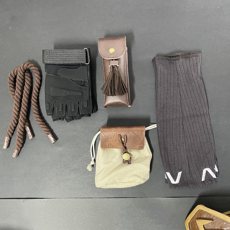 Pu&ograve; includere: Un assortimento di accessori tra cui una corda marrone, guanti senza dita neri, un astuccio in pelle marrone con nappina, una piccola borsa con coulisse beige e marrone e scaldamuscoli a coste grigio scuro con scritte bianche.