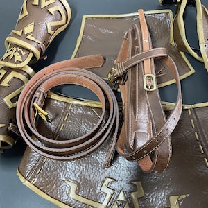 Pu&ograve; includere: Una collezione di pezzi di armatura in pelle marrone, tra cui una cintura con fibbia dorata, un corpetto e protezioni per le braccia. L'armatura presenta cuciture decorative e disegni in rilievo, che suggeriscono un tema medievale o fantasy.