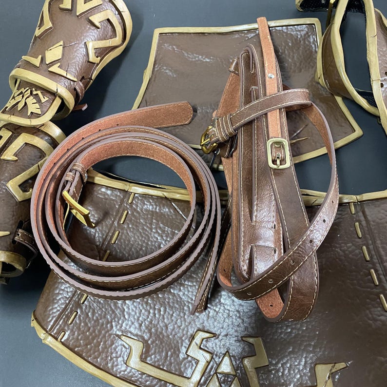 Pu&ograve; includere: Una collezione di pezzi di armatura in pelle marrone, tra cui una cintura con fibbia dorata, un corpetto e protezioni per le braccia. L'armatura presenta cuciture decorative e disegni in rilievo, che suggeriscono un tema medievale o fantasy.