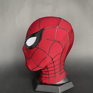 May include: A red and black Spider-Man mask, featuring a textured pattern and black web detailing. The mask has white mesh eye coverings and is displayed on a black stand. The mask is a cosplay item.