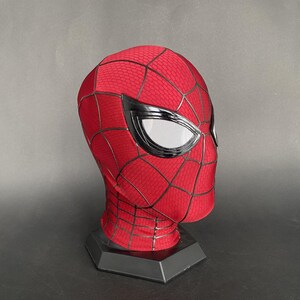 May include: A red and black Spider-Man mask with white mesh eye coverings. The mask has a web pattern and is displayed on a black stand. The mask is made of a flexible material.