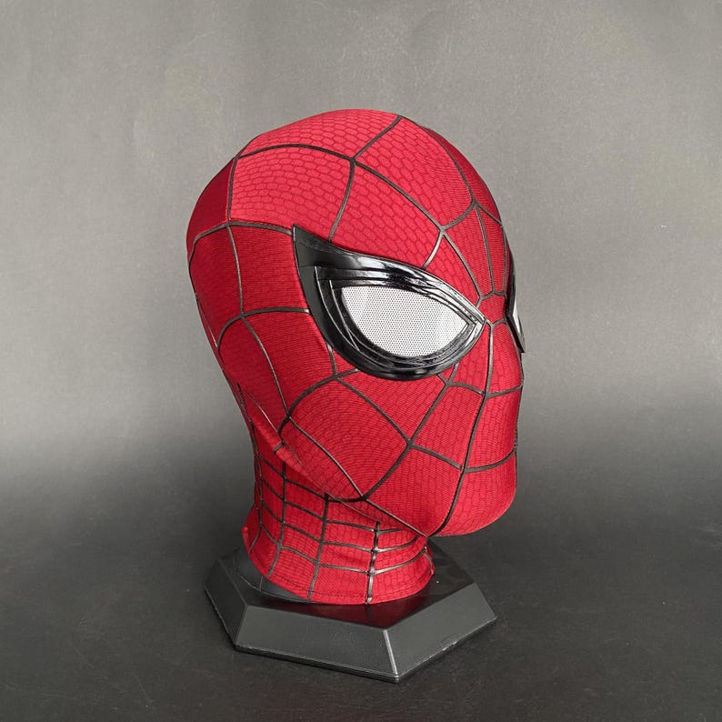 May include: A red and black Spider-Man mask with white mesh eye coverings. The mask has a web pattern and is displayed on a black stand. The mask is made of a flexible material.