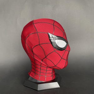 May include: A red and black Spider-Man mask with white eye lenses. The mask is displayed on a black stand. The mask has a webbed pattern and is made of a textured material.