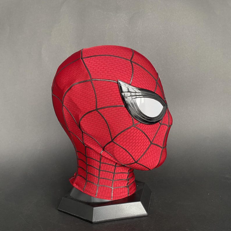 May include: A red and black Spider-Man mask with white eye lenses. The mask is displayed on a black stand. The mask has a webbed pattern and is made of a textured material.