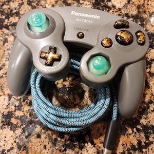 May include: A gray Panasonic SH-TGC10 controller with gold-colored buttons and a blue and black braided cord. The controller has a start/pause button and a cross-shaped button. The analog sticks are light green.