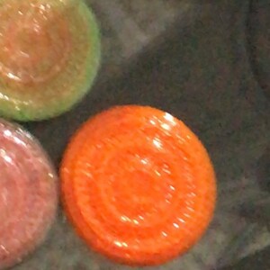 May include: A close-up of a round, orange scented candle with a decorative design. The candle is sitting on a dark surface.
