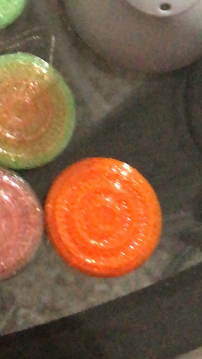May include: A close-up of a round, orange scented candle with a decorative design. The candle is sitting on a dark surface.