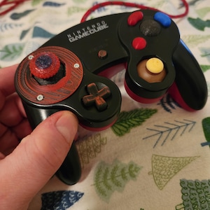 May include: Black Nintendo GameCube controller with red and blue buttons. The controller has a red and black design with the words "NINTENDO GAMECUBE" printed on the top. The controller is connected to a red cord.