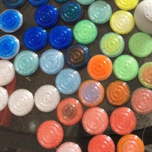 May include: A collection of round, glittery, colourful tiles in various shades of blue, green, orange, pink, and white. The tiles are arranged in a circular pattern.