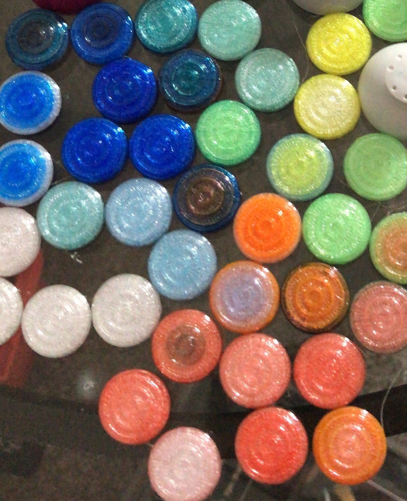 May include: A collection of round, glittery, colourful tiles in various shades of blue, green, orange, pink, and white. The tiles are arranged in a circular pattern.