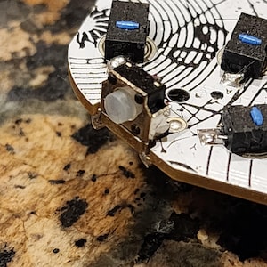 May include: Close-up of a circuit board with several black and silver buttons. The buttons have small blue accents. The board has a white surface with a black circular pattern. The background is a speckled brown and tan.