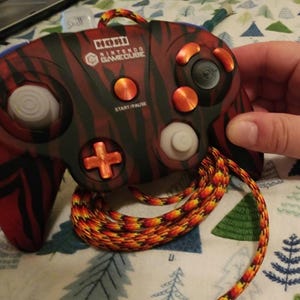 Peut inclure: Manette Hori Nintendo GameCube avec un motif rayé rouge et noir. La manette a des boutons orange et un stick analogique blanc. Le cordon est orange, jaune et rouge. Le texte "HORI Nintendo GameCube" est visible.