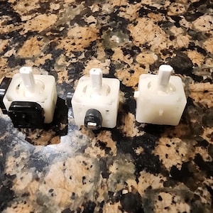 May include: Three white plastic rotary switches with black knobs and a black base. The switches are arranged in a row on a brown and white speckled surface.