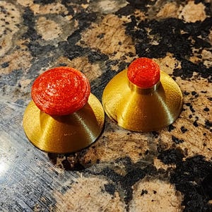 May include: Two decorative bottle stoppers. Each stopper features a gold-colored base with a red, textured, spherical top. The stoppers are displayed on a speckled, brown and gray surface.