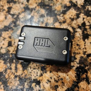 May include: A black rectangular electronic device with the letters "HHL" embossed on the top. The device has rounded corners, small silver screws, and a series of white vertical lines on one side.