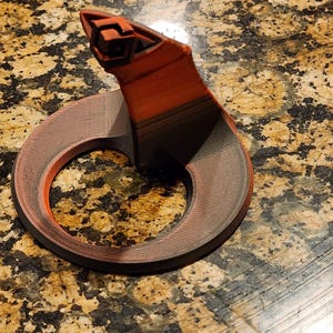 May include: A 3D printed, red and black, snake-shaped phone stand. The stand has a circular base and a raised head with a geometric design.