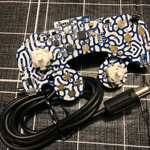 May include: A disassembled electronic device with a blue and white patterned circuit board. The board has various components, including white joysticks, and is connected to a black cable with a connector. The background is a black and white checkered pattern.