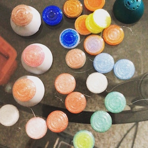 May include: Assortment of colourful round buttons with glitter and various textures. Some buttons are stacked on top of each other. The buttons are on a glass surface.