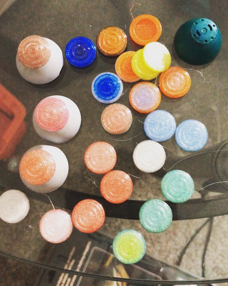 May include: Assortment of colourful round buttons with glitter and various textures. Some buttons are stacked on top of each other. The buttons are on a glass surface.