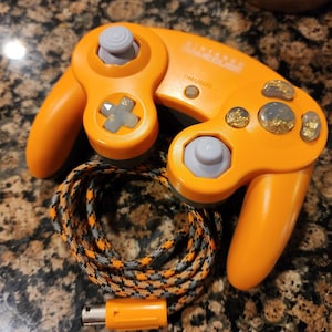 May include: Orange Nintendo GameCube controller with gray buttons and a matching orange cable. The controller has a distinctive design with a joystick, directional pad, and multiple buttons. The cable is black, orange, and gray.