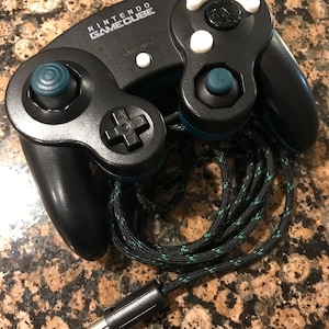 May include: A black Nintendo GameCube controller with a blue and green braided cord. The controller has white buttons and a black and teal circular button.
