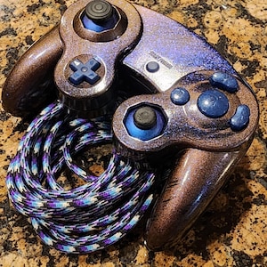 May include: A purple and brown GameCube controller with a black and white braided cord. The controller has a blue and black button layout and a sparkly finish.