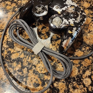 May include: Two black video game controllers with silver and gold leaf accents. The controllers have a gray and black braided cable. The controllers are connected by a gray velcro strap.