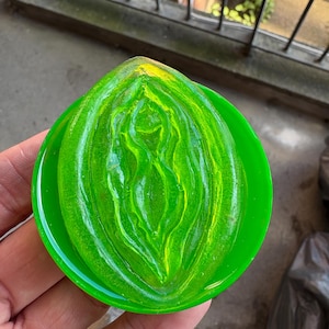 May include: A green, abstract sculpture with yellow highlights, resembling a stylized form, set within a circular green base. The artwork is held in a hand, showcasing the vibrant colours and unique design.