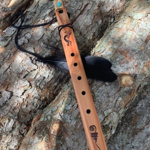May include: A wooden flute with a turquoise stone at the top, a black cord, and a black feather. The flute has a carved design of a Native American dancer and a swirl pattern.