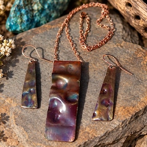 May include: A jewelry set consisting of a rectangular pendant on a copper chain and two matching trapezoidal drop earrings. Each piece features a hammered, iridescent finish with deep purple, blue, and bronze tones, suspended from copper hooks.