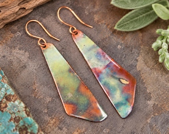 May include: A pair of long, flat, trapezoidal dangle earrings featuring a heat-treated finish with blended patches of copper, teal, green, and yellow. Each earring hangs from a simple copper wire hook, showcasing a smooth, metallic surface.