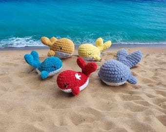 May include: A collection of six crocheted whale plushies in various colors, including blue, yellow, red, and gray, resting on a sandy beach. Each whale has a unique design, with white accents and small, closed eyes. The ocean is visible in the background.