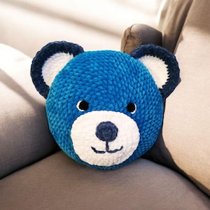 Op de afbeelding: Een blauw gebreid kussen in de vorm van een teddybeer met witte gezichtskenmerken. Het kussen ligt op een grijze bank.