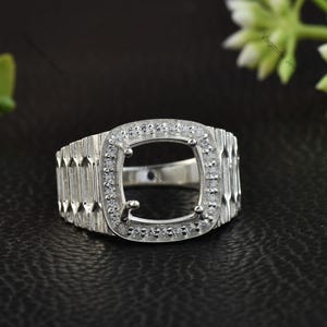 May include: A silver ring with a square center setting, surrounded by small, clear stones. The band features a textured, ridged design. The ring is set against a dark background, highlighting its metallic shine.