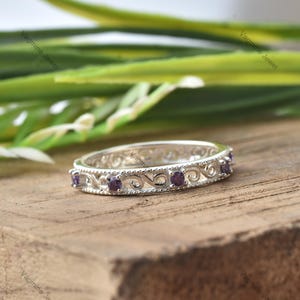 May include: A silver band ring with a delicate, ornate design featuring small purple gemstones. The ring is set against a wooden background.
