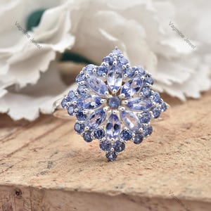 Könnte beinhalten: Ein silberner Ring mit einem blauen Edelstein-Blumenmuster. Der Ring ist mit vielen kleinen blauen Edelsteinen besetzt, die ein zartes und filigranes Blumenmuster bilden.