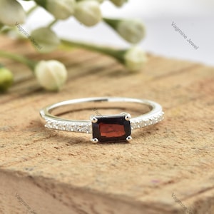 Könnte beinhalten: Ein silberner Ring mit einem rechteckigen roten Edelstein und einer Reihe kleiner, klarer Edelsteine um das Band.