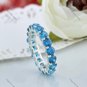 Könnte beinhalten: Ein silberner Bandring mit einer Reihe blauer Edelsteine. Der Ring ist mit runden, facettierten Steinen besetzt, die in einem durchgehenden Kreis angeordnet sind.