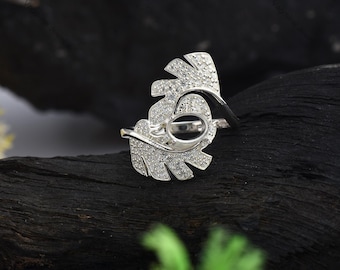 Puede incluir: Un anillo de plata con un diseño de hoja, que presenta una piedra transparente en el centro. El anillo está colocado sobre un fondo oscuro.
