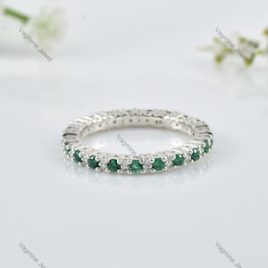 May include: A silver ring featuring a continuous row of small, round, emerald green gemstones and interspersed with small, clear gemstones. The ring is set against a white background, with the brand name "Vagmine Jewel" in the corners.