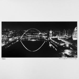 May include: A black and white photograph of the Millennium Bridge in Newcastle upon Tyne, England at night. The bridge is lit up and the reflection of the lights can be seen in the water.