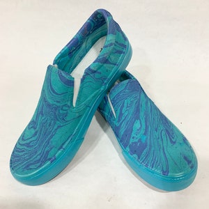 May include: A pair of slip-on shoes with a marbled design in shades of teal and blue. The shoes have a solid teal rubber sole and a white interior. The pattern resembles flowing water or paint swirls.