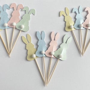 May include: Bunny-shaped cupcake toppers in pastel shades of yellow, blue, pink, and green. Each bunny has a white pom-pom tail and is attached to a wooden skewer. Ideal for Easter or spring celebrations. The toppers are made of paper.