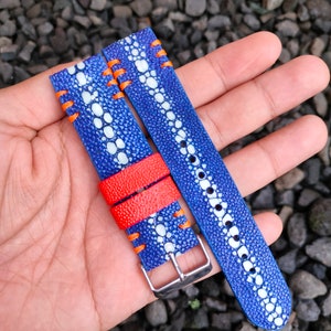 May include: A blue and white patterned leather watch strap with orange stitching. The strap is folded in half and held in a hand.