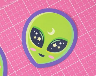 May include: Three green alien face stickers with purple outlines. Each alien has a crescent moon, stars, and black eyes. The stickers are on a pink cutting mat.