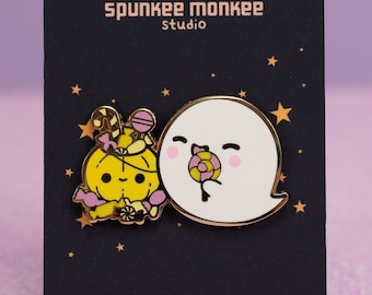 Op de afbeelding: Twee emaillepins met een spook en een pompoen die snoep vasthouden. De pins bevinden zich op een zwarte kaart met de tekst "Speckles Trick or Treat" en "Spunkee Monkee Studio" in gouden letters. De kaart vermeldt ook "emaillepins van Britlos."