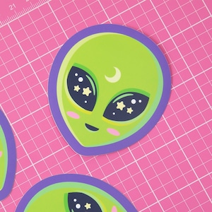 Peut inclure: Trois autocollants représentant des visages d'extraterrestres verts avec des contours violets. Chaque extraterrestre a une lune croissante, des étoiles et des yeux noirs. Les autocollants sont sur un tapis de coupe rose.