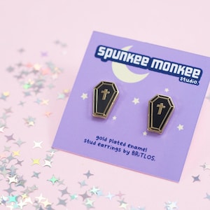 May include: Gold plated enamel coffin stud earrings by BRITLOS. The earrings are black with a gold cross on each coffin. The earrings are on a purple card with the text "Spunkee Monkee Studio" and "gold plated enamel stud earrings by BRITLOS."