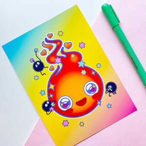May include: A colorful art print featuring a cartoon fire demon with large eyes, surrounded by stars and hearts. Two small, dark, fluffy creatures are also present. A green pen rests on the pink and white surface.