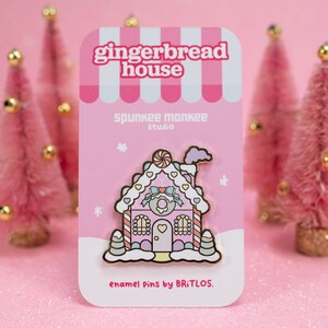 May include: A pink gingerbread house enamel pin with white frosting, candy canes, and a pink bow. The pin is on a pink card with the text "gingerbread house", "spunkee monkee studio", and "enamel pins by BRITLOS."